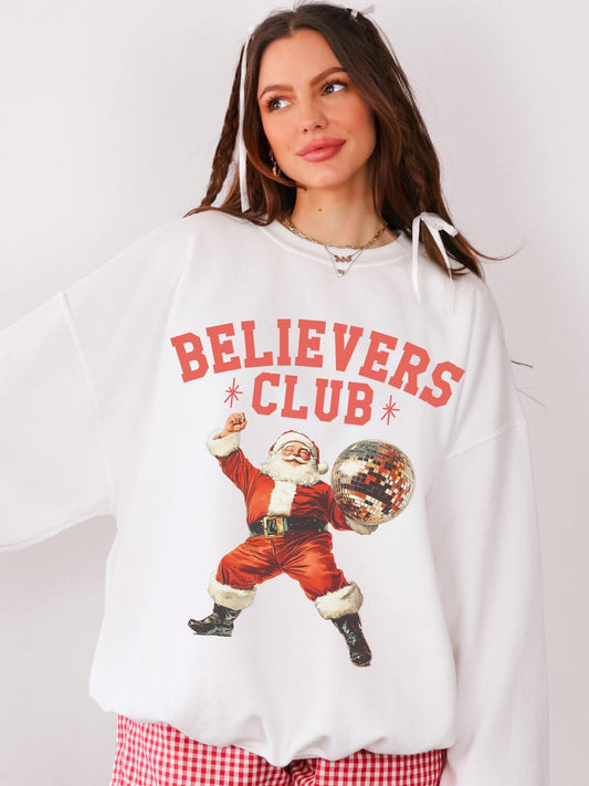 Model is wearing red checker boxer shorts and an oversized white crewneck sweatshirt featuring a Christmas graphic. The Christmas graphic is of a retro dancing Santa holding a disco ball with the words, believers club.