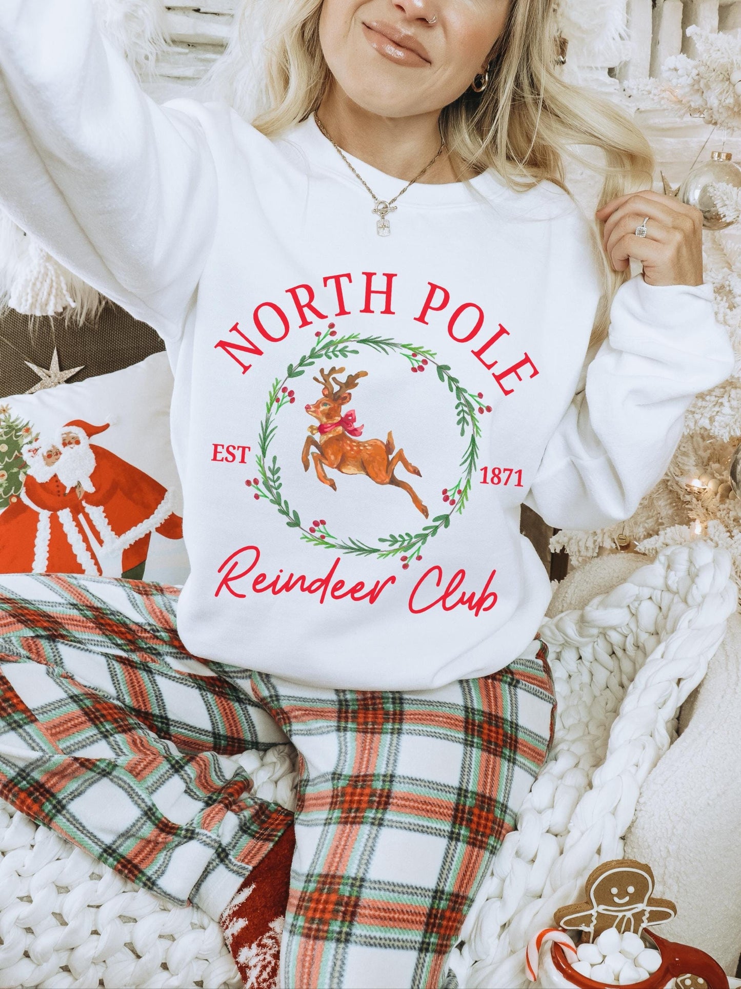 Model surrounded by a Christmas setup while wearing an oversized crewneck sweatshirt featuring a Christmas graphic. The holiday graphic is of a reindeer with a red bow surrounded by a holly wreath surrounded by the words north pole reindeer club