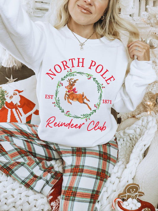 Model surrounded by a Christmas setup while wearing an oversized crewneck sweatshirt featuring a Christmas graphic. The holiday graphic is of a reindeer with a red bow surrounded by a holly wreath surrounded by the words north pole reindeer club